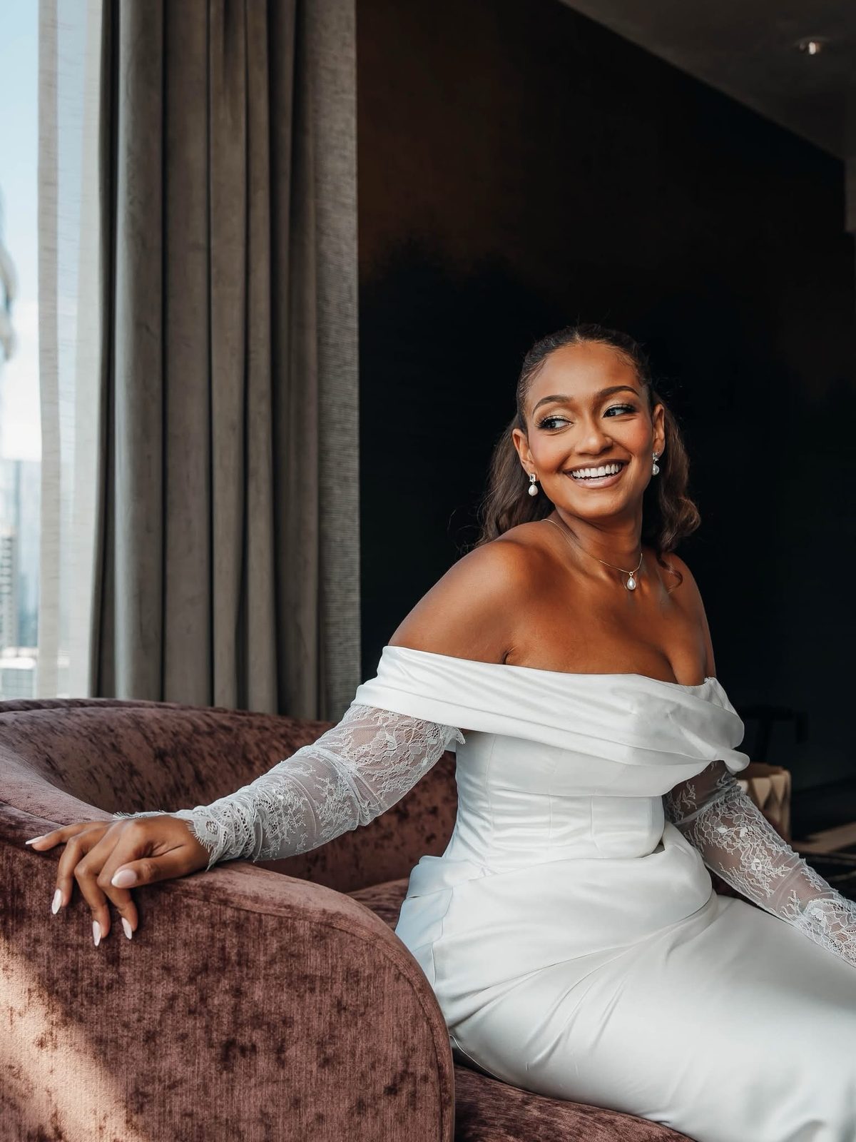 Bride in an off-shoulder lace-sleeve gown, smiling on a velvet chair