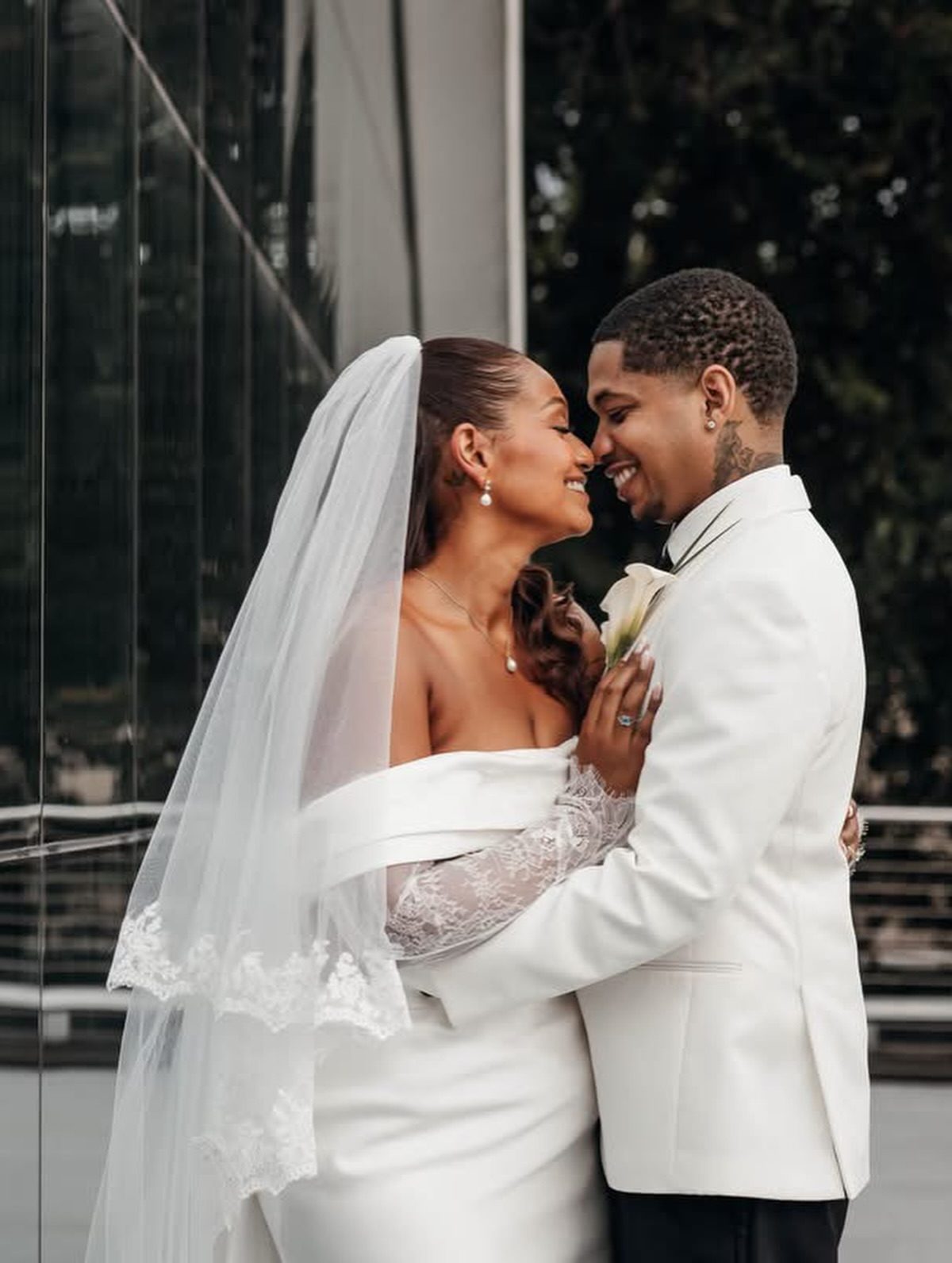 Bride and groom in white, faces close, beside a city window